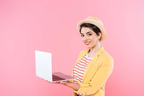 Mujer de raza bastante mixta en sombrero de paja usando el ordenador portátil mientras sonríe a la cámara aislada en rosa - foto de stock