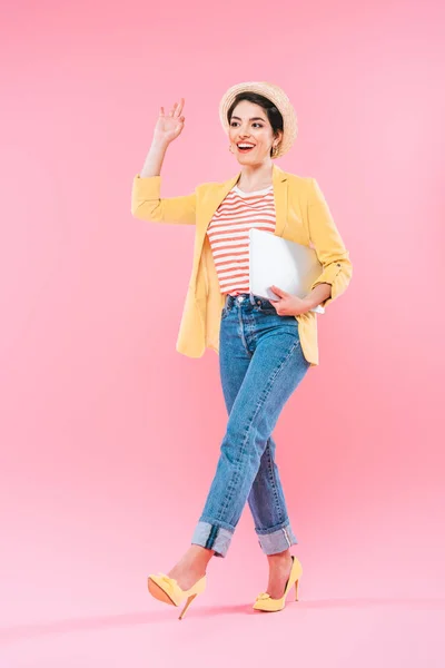 Aufgeregte Mischlingsfrau geht mit Laptop und winkender Hand auf rosa Hintergrund — Stockfoto