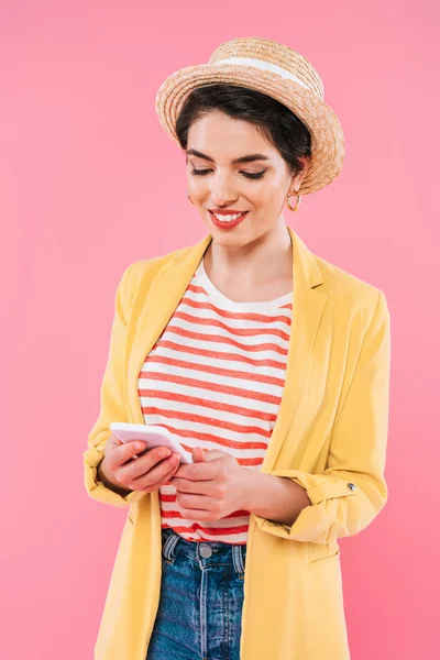 Souriant mixte femme en vêtements lumineux et chapeau de paille à l'aide d'un smartphone isolé sur rose — Photo de stock