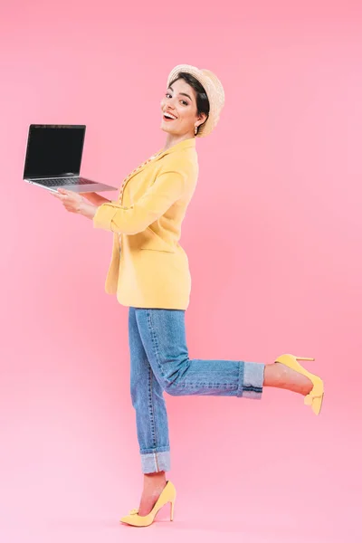 Alegre mujer de raza mixta en ropa brillante y sombrero de paja posando en la cámara con el ordenador portátil sobre fondo rosa - foto de stock