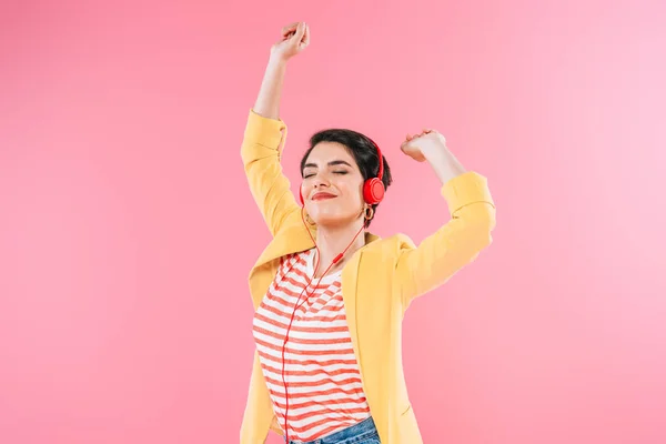 Emocionada mujer de raza mixta escuchando música en auriculares y bailando aislada en rosa - foto de stock