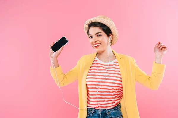 Mujer de raza bastante mixta escuchando música en auriculares y sosteniendo el teléfono inteligente con pantalla en blanco aislado en rosa - foto de stock
