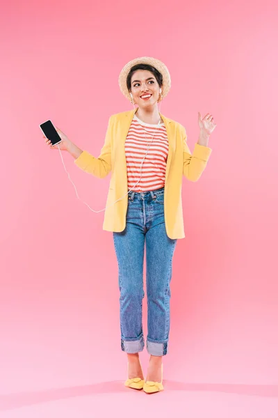 Atractiva mujer de raza mixta escuchando música en auriculares y sosteniendo el teléfono inteligente con pantalla en blanco sobre fondo rosa - foto de stock