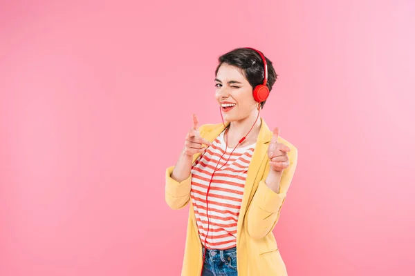 Alegre mujer de raza mixta escuchando música en auriculares, señalando con los dedos y guiñando un ojo a la cámara aislada en rosa - foto de stock