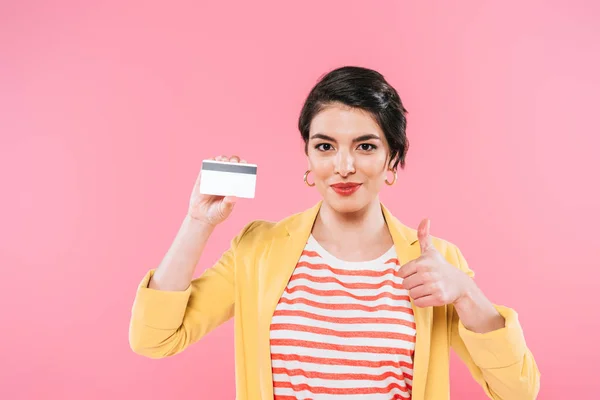 Hermosa mujer de raza mixta sosteniendo tarjeta de crédito y mostrando el pulgar hacia arriba aislado en rosa - foto de stock