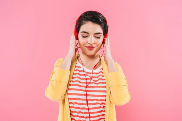 Mulher de raça mista sonhadora ouvindo música em fones de ouvido com olhos fechados isolados em rosa — Fotografia de Stock