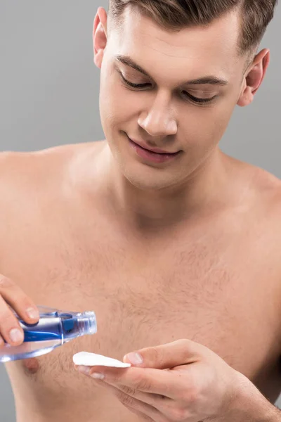 Smiling nude young man applying lotion on cotton pad isolated on grey — Stock Photo