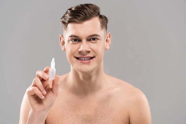 Vista frontal del joven desnudo sonriente sosteniendo gotas para los ojos y mirando a la cámara aislada en gris - foto de stock