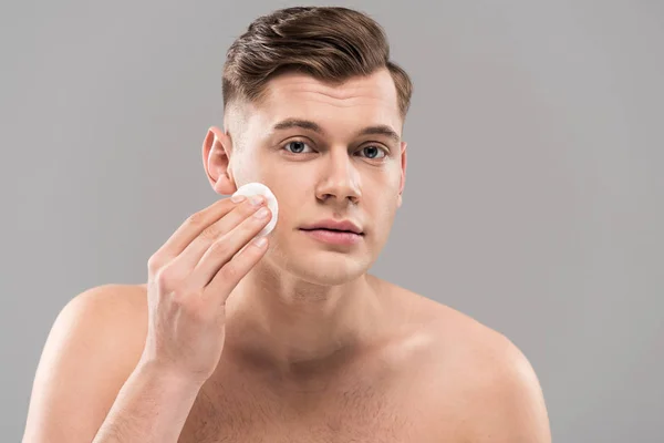 Bel giovane uomo pulire il viso con batuffolo di cotone isolato su grigio — Foto stock