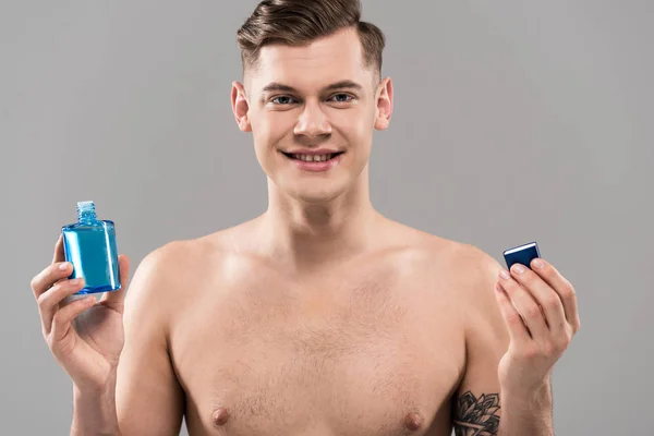 Vorderansicht eines lächelnden nackten jungen Mannes mit einer Lotionflasche isoliert auf grau — Stockfoto