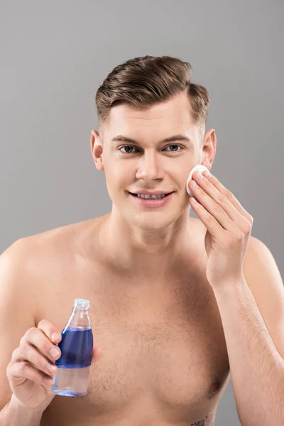 Vista frontal de un joven desnudo sonriente sosteniendo una botella de loción y limpiando la cara con una almohadilla de algodón aislada en gris - foto de stock