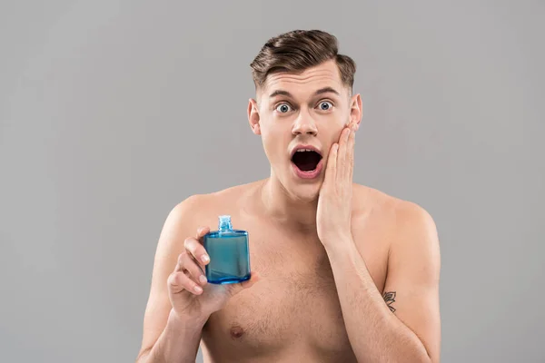 Вид спереду шокованого голого молодого чоловіка, що тримає пляшку і наносить лосьйон на обличчя ізольовано на сірий — стокове фото