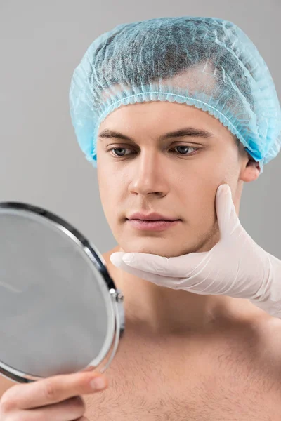 Partial view of cosmetologist in latex glove and young man in medical cap isolated on grey — Stock Photo