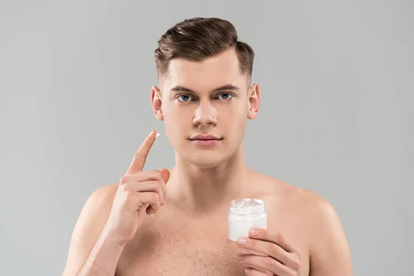 Vorderseite des nackten jungen Mannes mit Kosmetikbehälter und Anwendung Gesichtscreme isoliert auf grau — Stockfoto