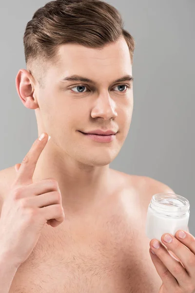 Голий молодий чоловік тримає косметичну тару і наносить крем для обличчя ізольовано на сірий — стокове фото