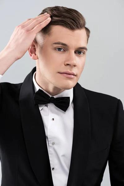 Smiling young man in formal wear with bow tie touching hair isolated on grey — Stock Photo