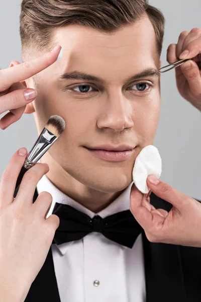 Vista recortada de los artistas de maquillaje haciendo maquillaje para el hombre en el desgaste formal sosteniendo vaso de cóctel aislado en gris - foto de stock