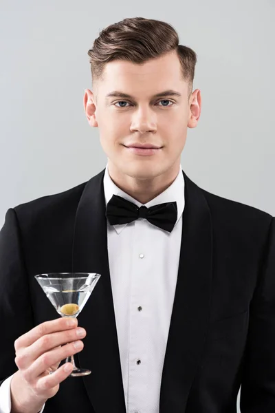 Front view of smiling young man in formal wear with bow tie holding glass of cocktail isolated on grey — Stock Photo