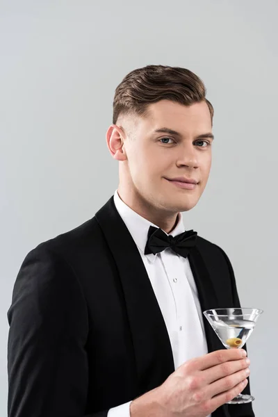 Joven sonriente en ropa formal con corbata de lazo sosteniendo vaso de cóctel aislado en gris — Stock Photo