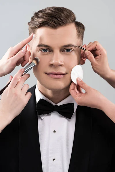 Обрізаний вид візажистів, які роблять макіяж для людини в офіційному одязі, тримаючи склянку коктейлю ізольовано на сірому — стокове фото