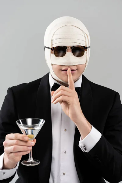 Vista frontal del hombre en ropa formal y gafas de sol con vendaje elástico en la cabeza sosteniendo vaso de cóctel y mostrando el signo de silencio aislado en gris - foto de stock