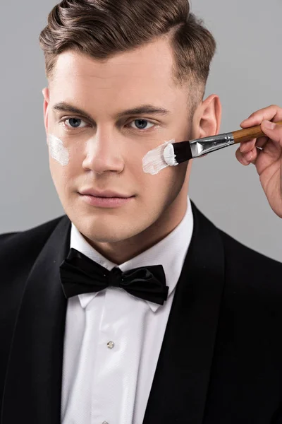 Cropped view of cosmetologist applying cream on face and man in formal wear isolated on grey — Stock Photo