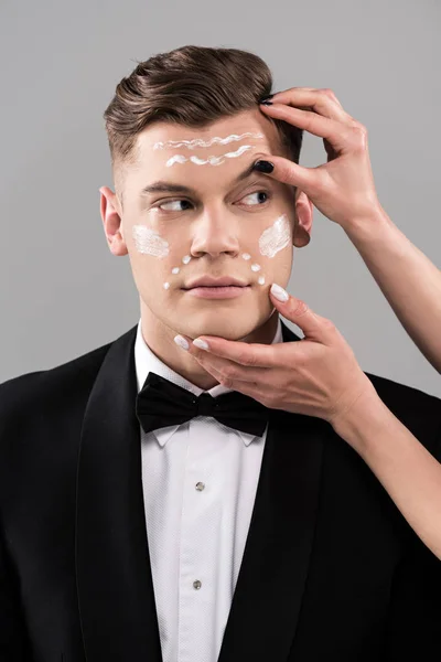 Vista parcial de cosmetólogos aplicando crema y hombre en ropa formal aislado en gris - foto de stock