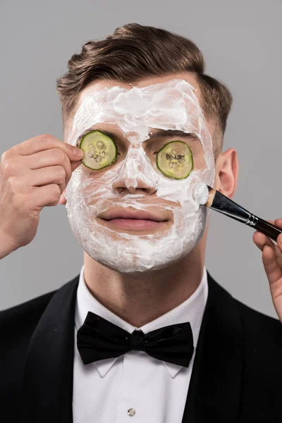 Partial view of cosmetologists applying cucumber mask and man in formal wear isolated on grey — Stock Photo