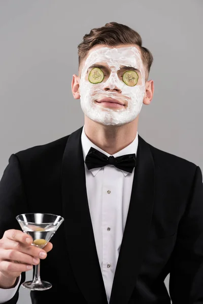 Jeune homme en tenue formelle avec masque facial concombre tenant verre de cocktail isolé sur gris — Photo de stock