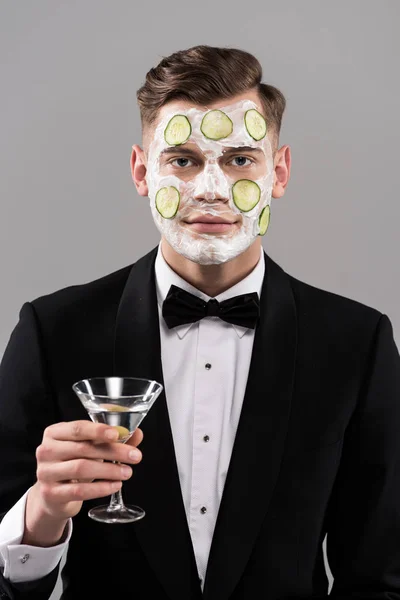 Jeune homme en tenue formelle avec masque facial concombre tenant verre de cocktail isolé sur gris — Photo de stock