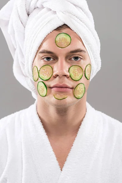 Front view of young man in bathrobe with cut cucumbers on face isolated on grey — Stock Photo