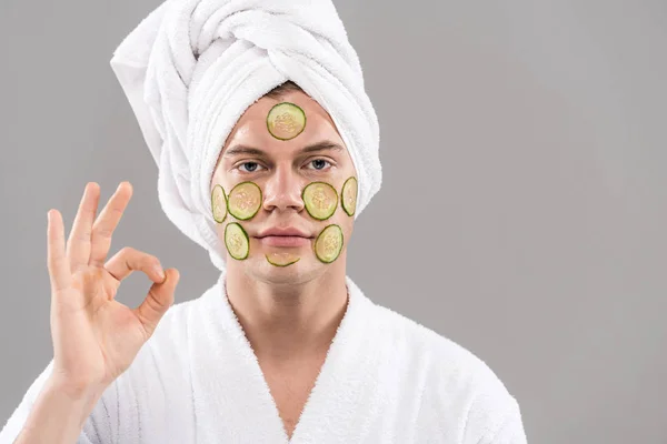 Vorderansicht des Mannes mit geschnittenen Gurken im Gesicht, die okay Zeichen isoliert auf grau zeigen — Stockfoto