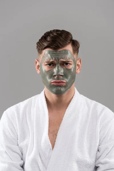 Front view of sad young man in bathrobe with clay mask isolated on grey — Stock Photo