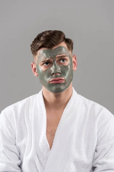 Front view of sad young man in bathrobe with clay mask isolated on grey — Stock Photo