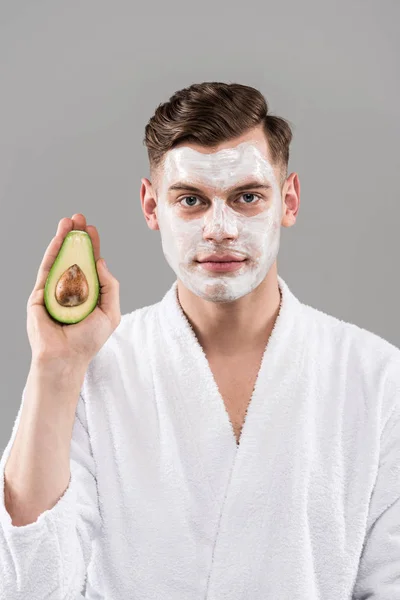 Вид спереди человека в халате с кремом на лице, держащего разрезанный авокадо изолированный на серый — стоковое фото