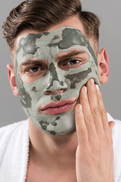 Unzufriedener junger Mann mit Tonmaske berührt Gesicht isoliert auf grau — Stockfoto