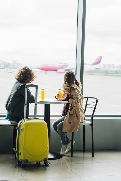Niños sentados en la mesa con jugo cerca de la maleta amarilla y mirando por la ventana en el avión - foto de stock