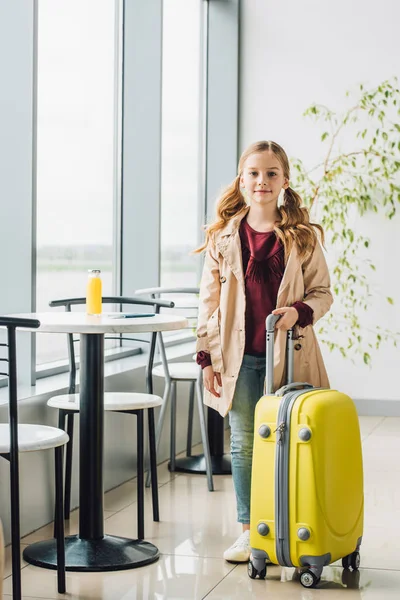 Full length view of preteen kid standing near table and chair with yellow suitcase in airport — Stock Photo