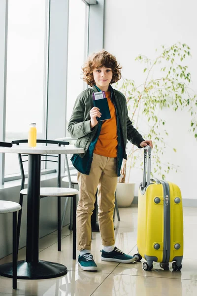 Vue pleine longueur du garçon debout avec valise jaune, tenant passeport et billet dans la salle d'attente à l'aéroport — Photo de stock