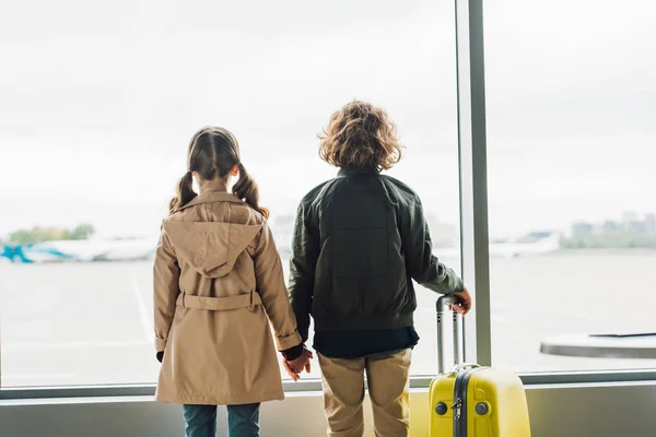 Вид сзади детей, держащихся за руки, стоящих у окна в зале ожидания в аэропорту — стоковое фото