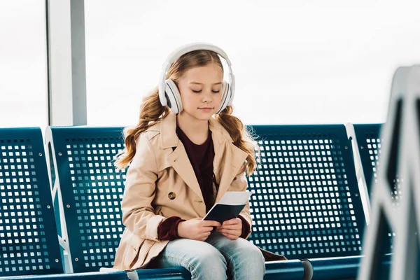 Preteen kid sitting on blue seat, holding passport and listening to music in waiting hall in airport — Stock Photo