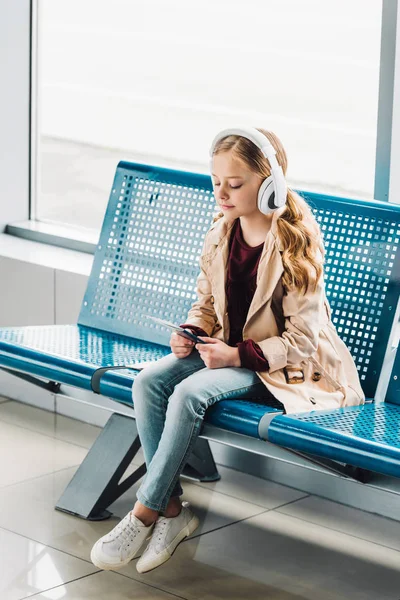 Vista completa del bambino preadolescente seduto sul sedile blu, in possesso di passaporto e ascolto di musica in sala d'attesa in aeroporto — Foto stock