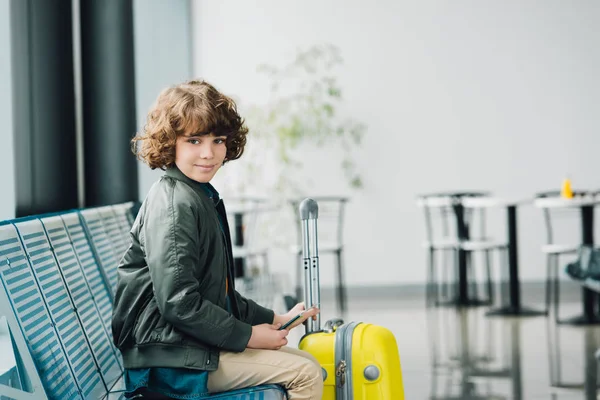 Niño sentado en el asiento azul, con pasaporte y maleta amarilla y mirando a la cámara en la sala de espera en el aeropuerto - foto de stock