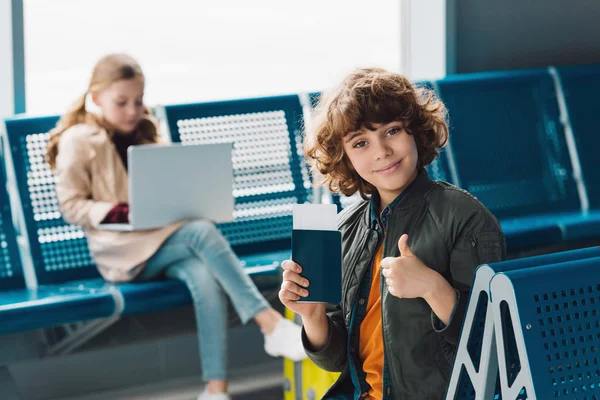 Selective focus on boy holding passport and showing thumb up while sitting on blue seat in waiting hall — Stock Photo