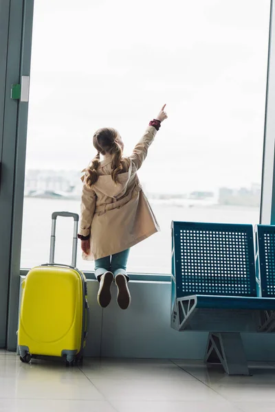 Vista trasera del niño preadolescente cerca de la maleta apuntando con el dedo a la ventana en la sala de espera en el aeropuerto - foto de stock
