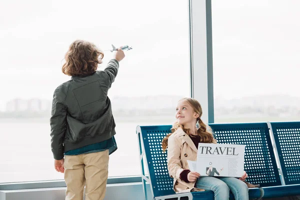 Cute preteen kid sitting in waiting hall with travel newspaper while boy playing with toy plane — Stock Photo