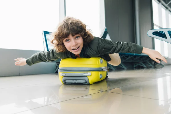 Niño preadolescente emocionado acostado en la maleta con las manos extendidas en la sala de salida del aeropuerto - foto de stock