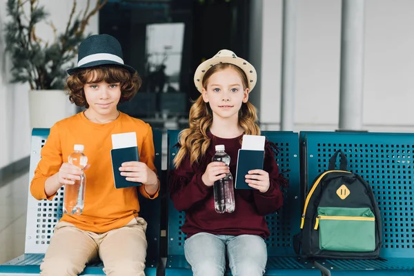 Niños preadolescentes lindos sentados en la sala de espera con botellas de agua, pasaportes y boletos de avión - foto de stock