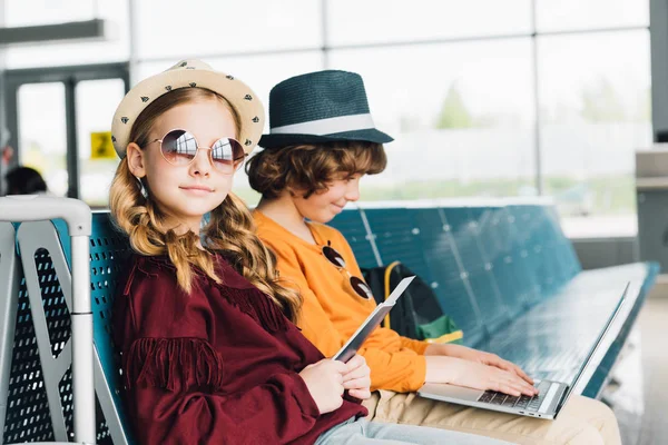 Cute preteen kids sitting in waiting hall with laptop and passport — Stock Photo