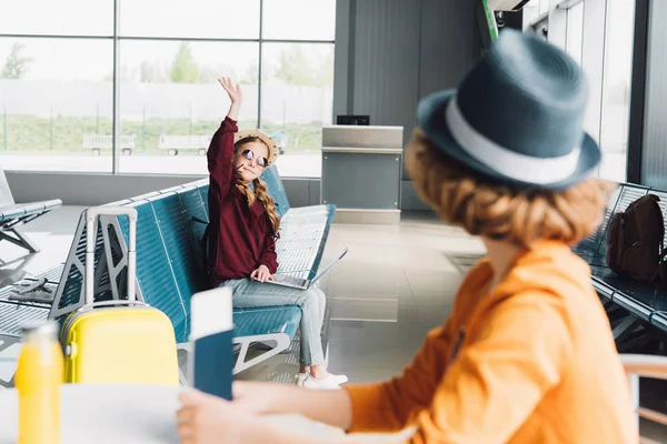 Niño preadolescente con portátil saludando a niño preadolescente con pasaporte y billete de avión en la sala de espera - foto de stock